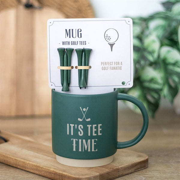A green mug with the text 'IT'S TEE TIME' printed on it, accompanied by an image of a golf club and golf tees, placed on a wooden surface with a plant in the background.