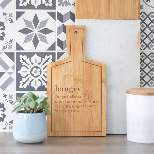 Wooden cutting board with 'hangry' definition text, potted plant, and white container against a patterned wall.
