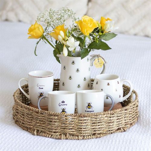 Set of white mugs with bee design on a woven tray with yellow flowers in a vase.