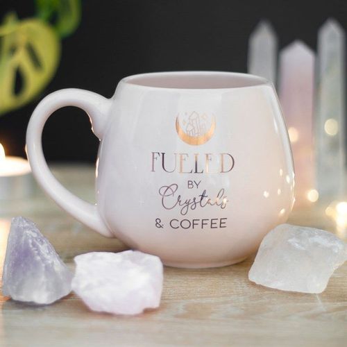White mug with 'Fueled by Crystals & Coffee' text on a table with crystals and blurred lights in the background.